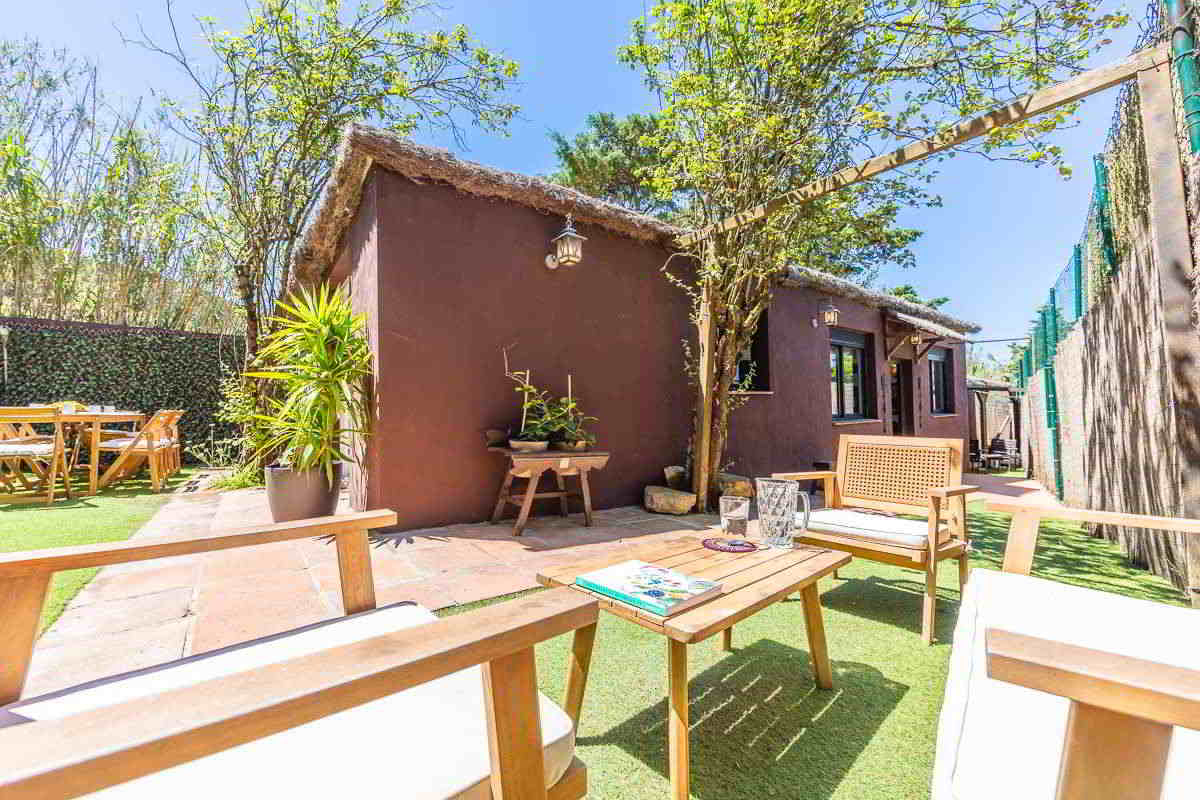 Detalle del porche y zona de descanso exterior de Villa Verde en Tarifa, con sillones y mesa de madera, rodeados de vegetación y muros color tierra.