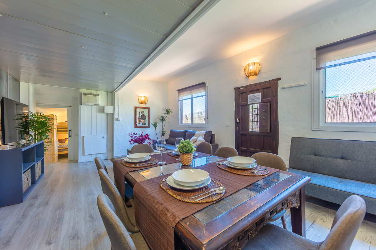 Comedor con mesa de madera preparada para cuatro comensales, sofá cama gris, puerta de entrada rústica de madera, ventanas con luz natural y vista a habitación con litera al fondo.
