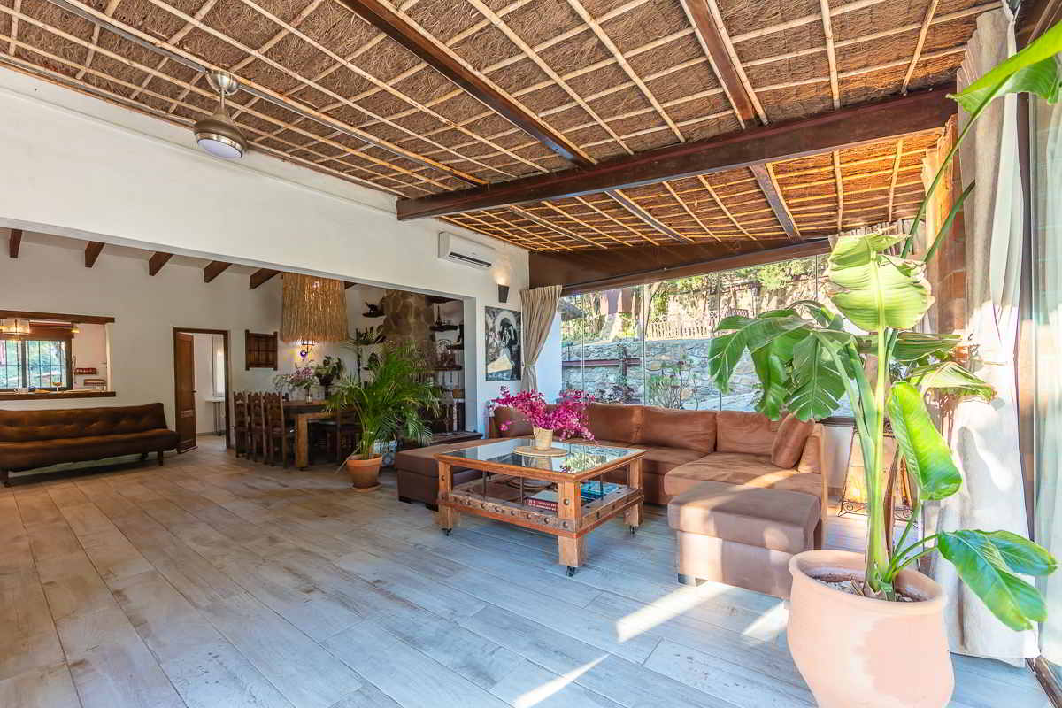 Espacio abierto que conecta el salón principal con el porche acristalado del chalet La Ahumada, con sofás de cuero, mesa de centro rústica, plantas decorativas y techo de cañizo.