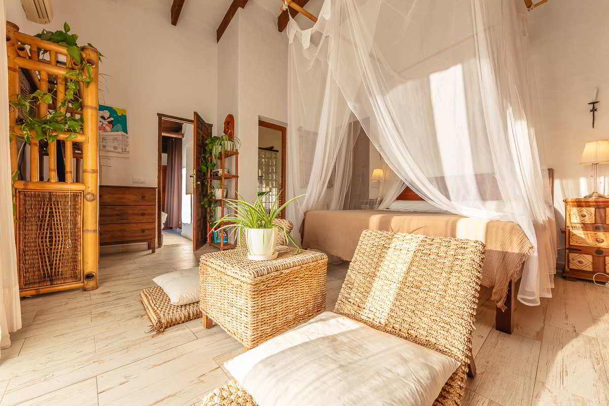 Interior del dormitorio principal del chalet La Ahumada con cama con dosel, sillas bajas de mimbre, mueble de bambú con plantas y decoración artesanal.