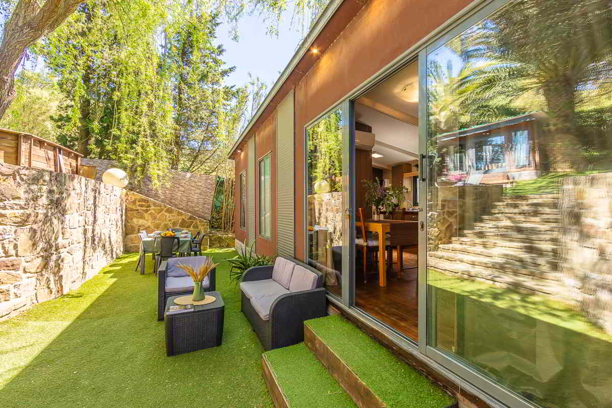 Zona exterior ajardinada de la casa Nepal en Villas La Ahumada, con césped artificial, sillones de ratán, mesa de comedor al fondo, muro de piedra y grandes ventanales que conectan con el interior de la vivienda