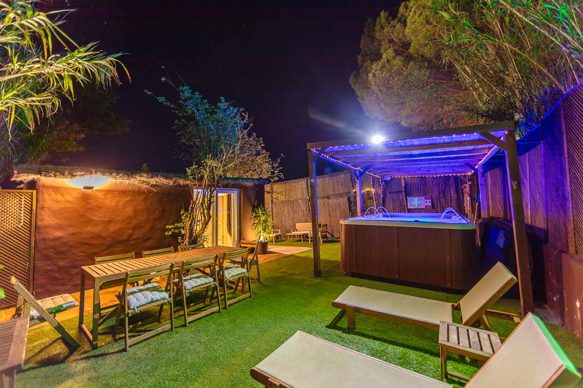 Vista nocturna de un jardín con césped artificial, tumbonas, mesa de comedor con sillas acolchadas, jacuzzi bajo pérgola iluminada y casita al fondo con puerta abierta