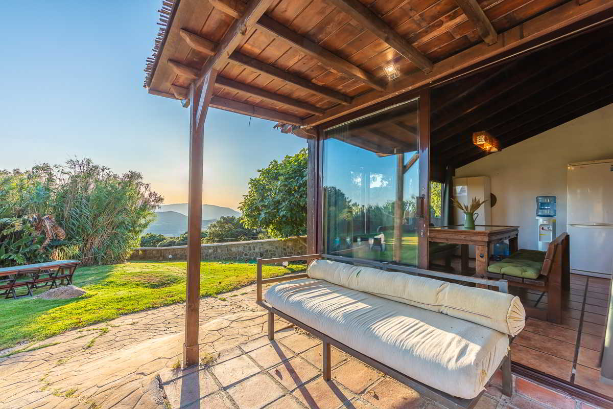 Porche cubierto de Villa Paraíso en Villas La Ahumada, Tarifa, con sofá acolchado, mesa de comedor interior y vistas al jardín y las montañas al atardecer.