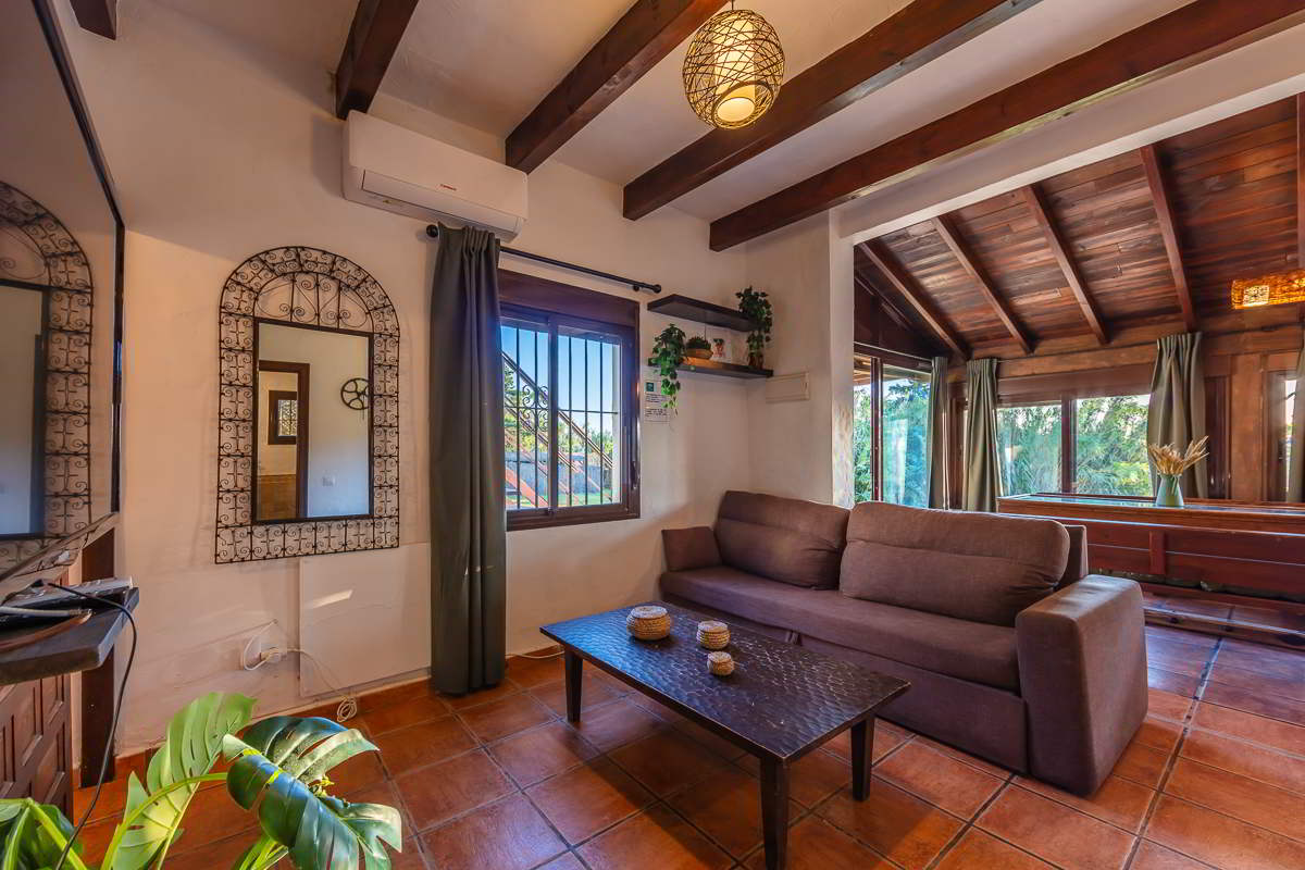 Salón rústico de Villa Paraíso, Villas La Ahumada, Tarifa, con sofá marrón, mesa de madera oscura, espejo decorativo con marco de forja, techo de vigas y ventanales con vistas al jardín.