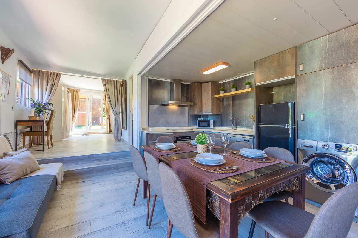 Vista de la cocina abierta de la Villa Verde con mobiliario en tonos cobre envejecido, mesa de comedor rústica con vajilla dispuesta, lavadora, nevera negra, plantas decorativas y fondo con salida a la terraza.