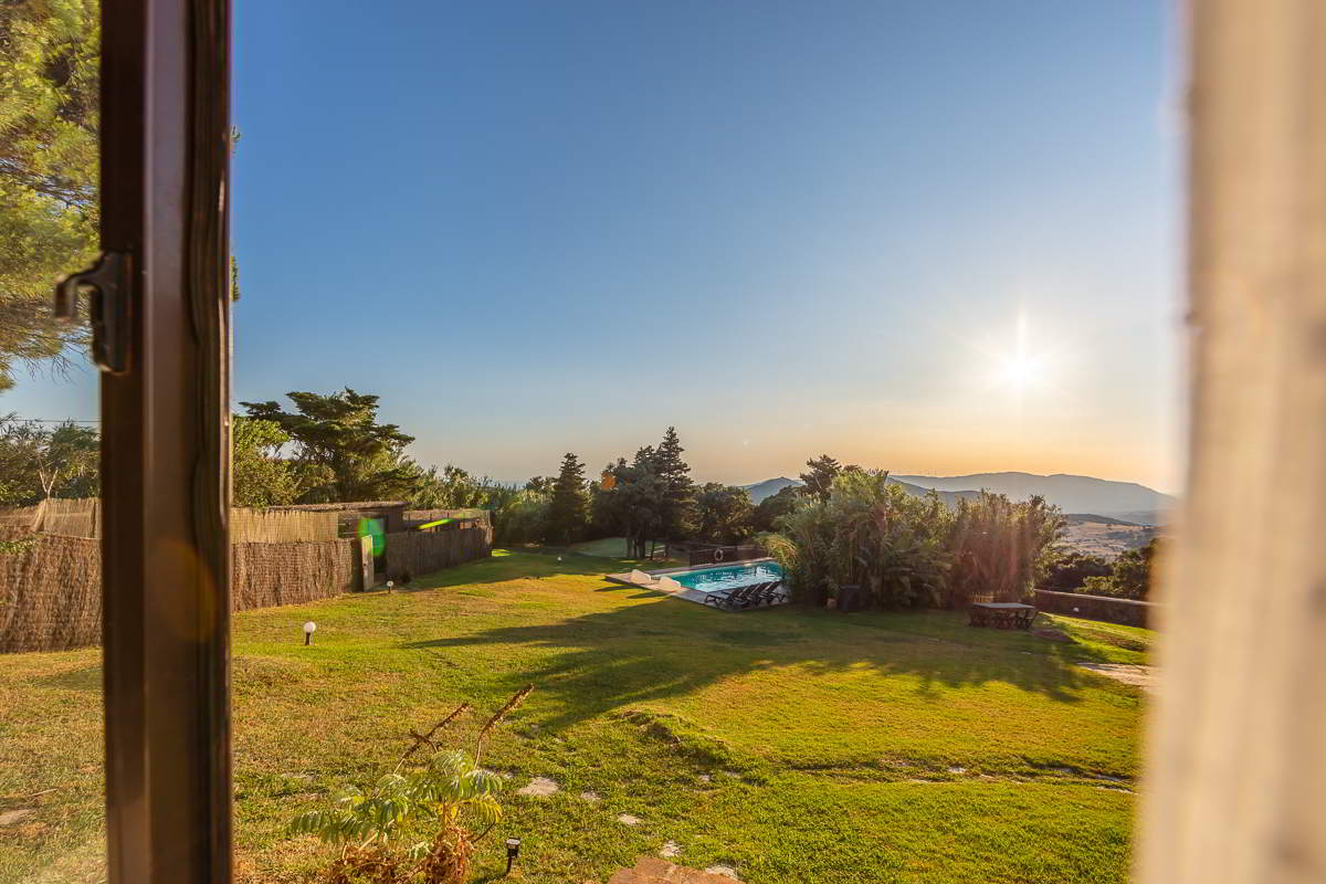 Vista desde una ventana del chalet La Ahumada al gran jardín con piscina y al fondo las montañas al atardecer.