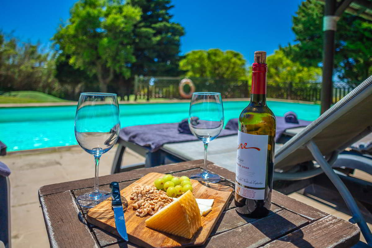 Mesa con vino, queso, uvas y nueces junto a tumbonas en la piscina del Chalet La Ahumada, rodeada de vegetación y luz de verano.