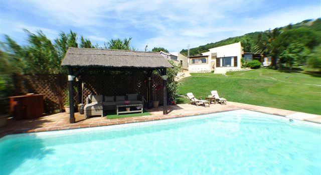 Piscina al aire libre con zona de descanso cubierta, rodeada de naturaleza y un amplio jardín con vistas al chalet.