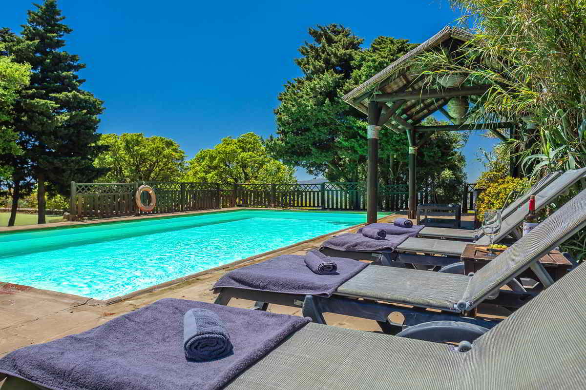 Piscina del Chalet La Ahumada con tumbonas, toallas, pérgola de madera y vegetación alrededor, bajo un cielo azul intenso.