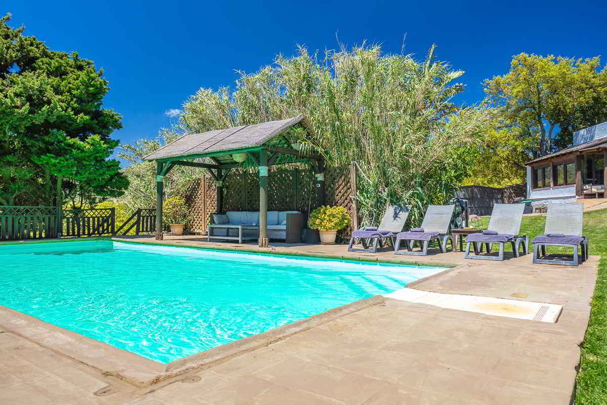 Vista general de la piscina del Chalet La Ahumada con tumbonas, pérgola de sombra y parte del edificio principal al fondo rodeado de vegetación.