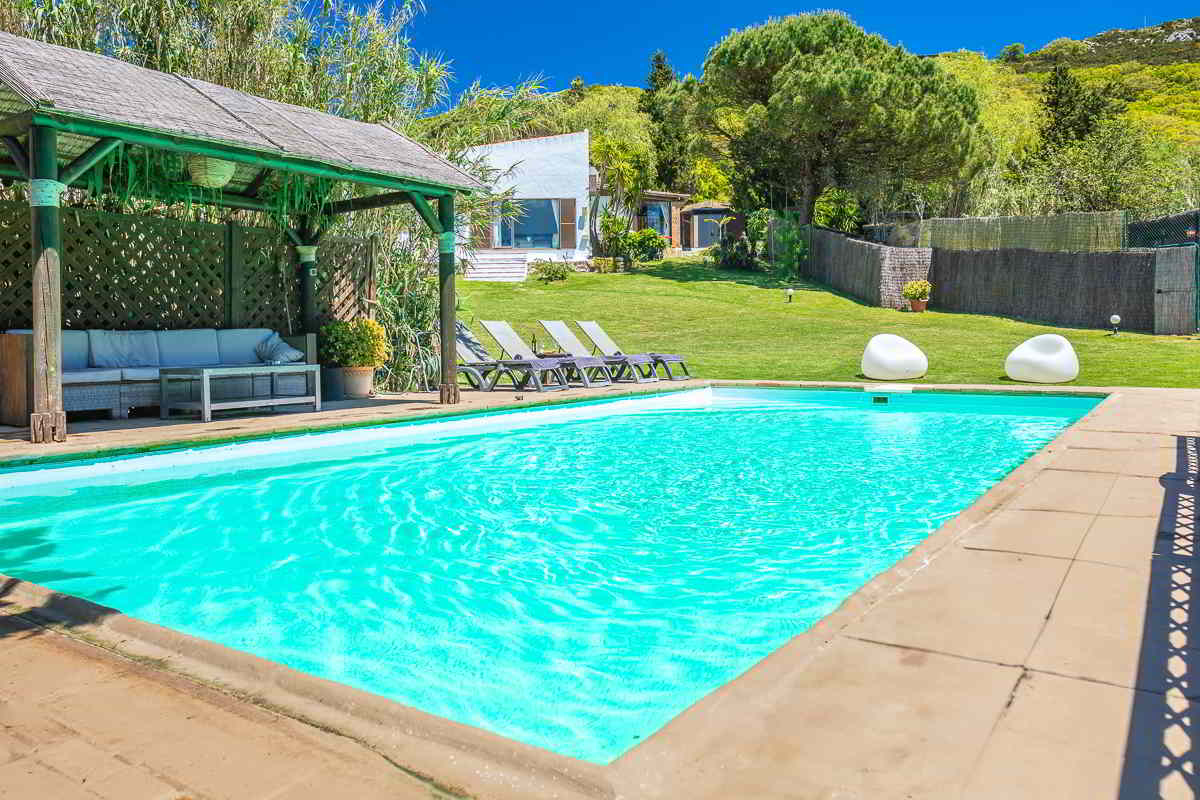 Piscina del Chalet La Ahumada con zona chill-out bajo pérgola de madera, tumbonas alineadas y edificio principal al fondo rodeado de césped.