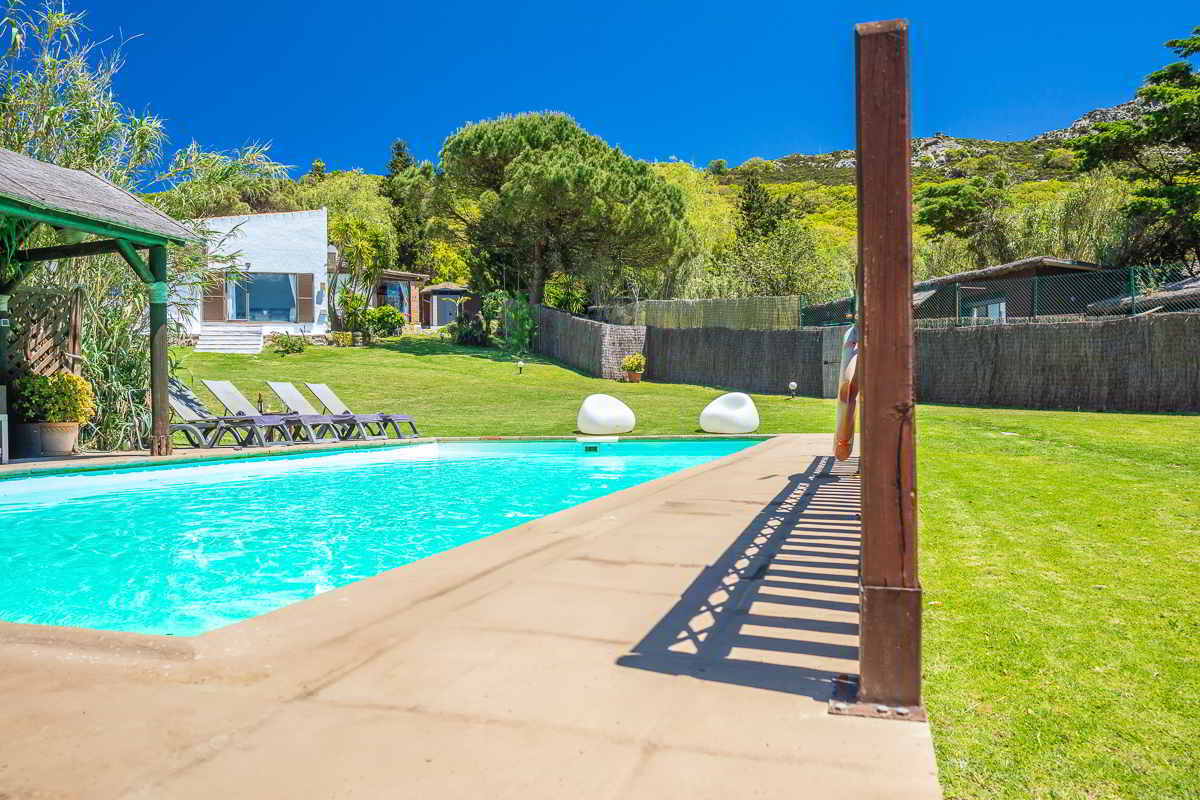 Piscina del Chalet La Ahumada con borde amplio, tumbonas, pérgola vegetal y vistas al edificio principal rodeado de jardín y vegetación.