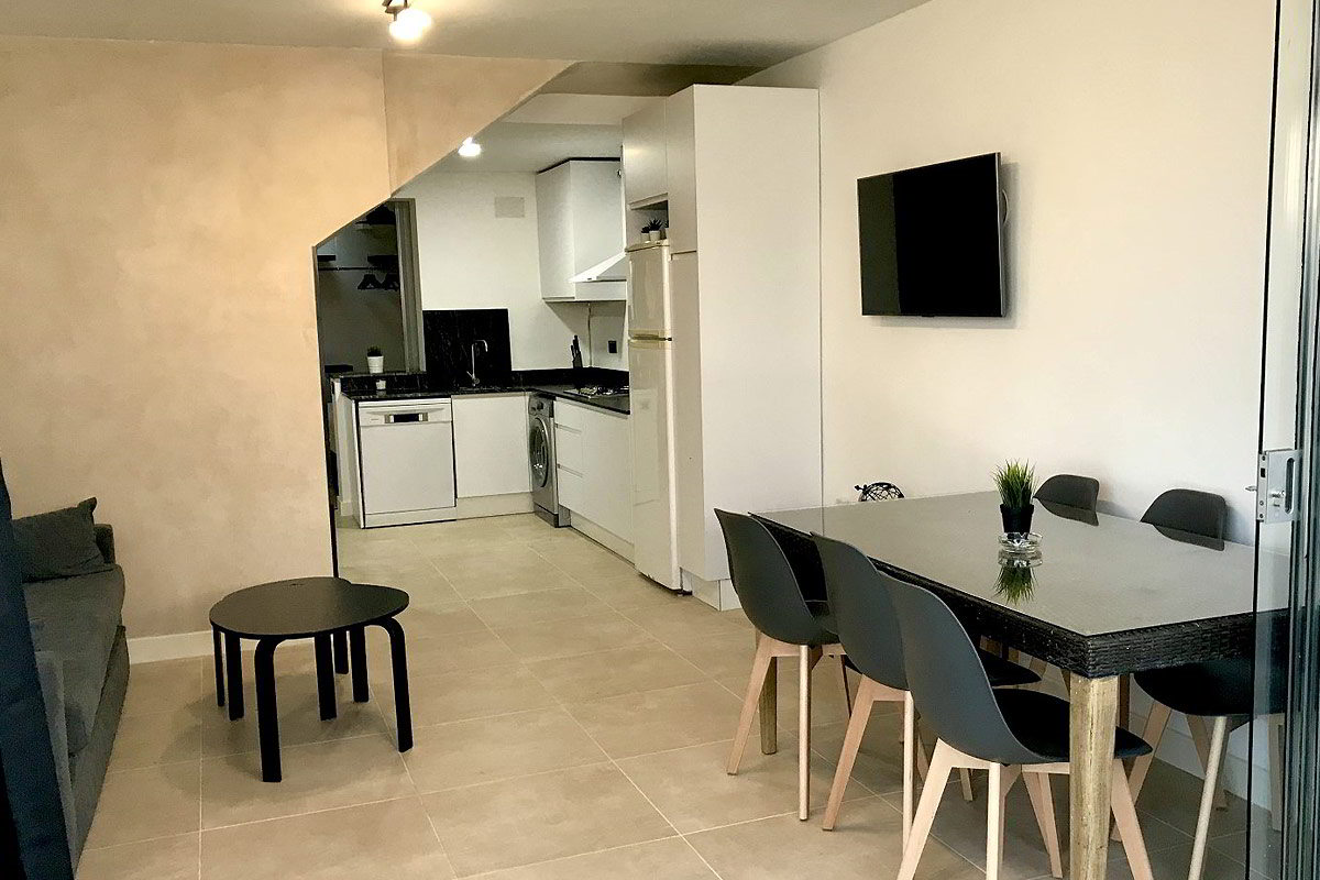 Salón moderno con comedor, mesa de cristal, sillas grises y televisor montado en la pared.