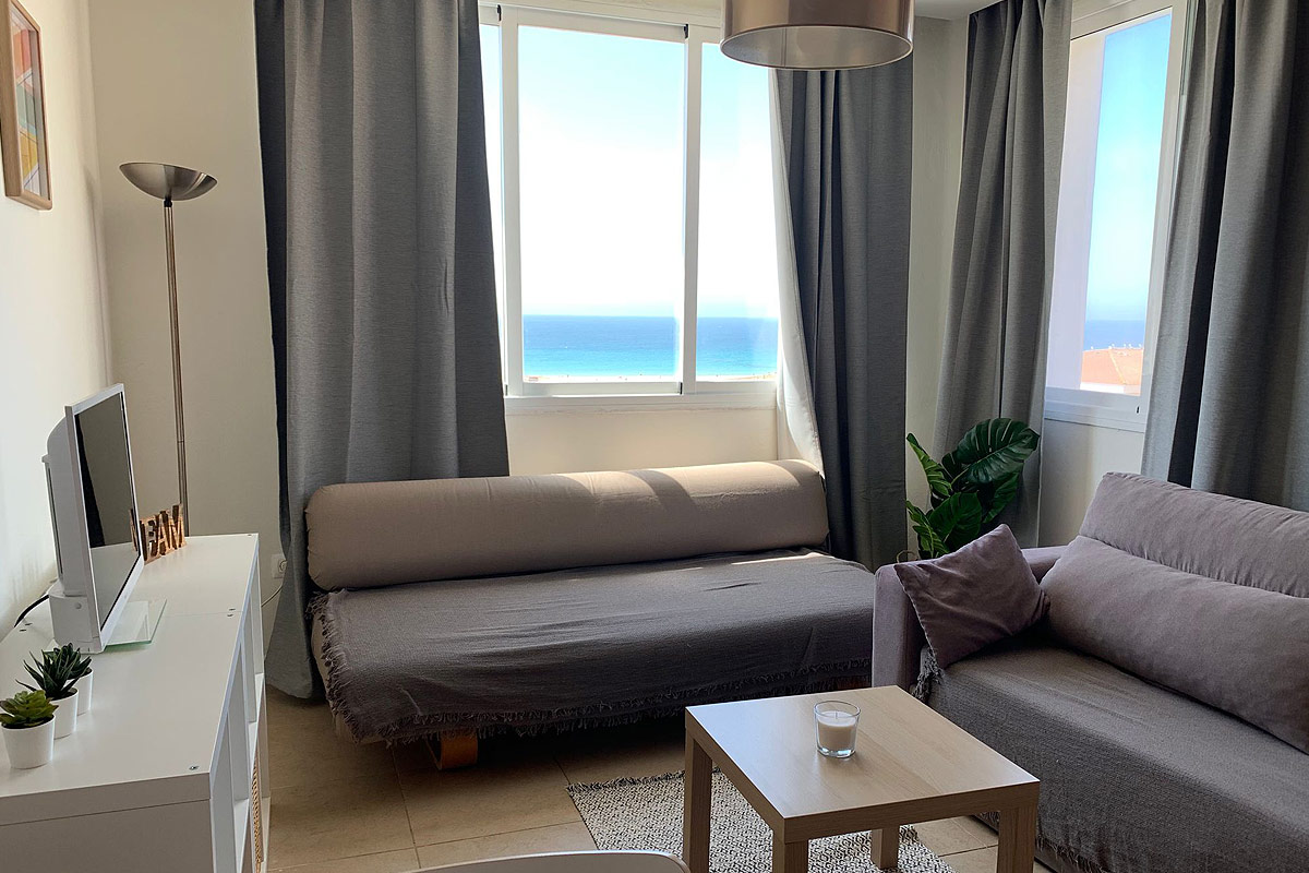 Salón con sofá gris, ventana grande y vistas al mar.