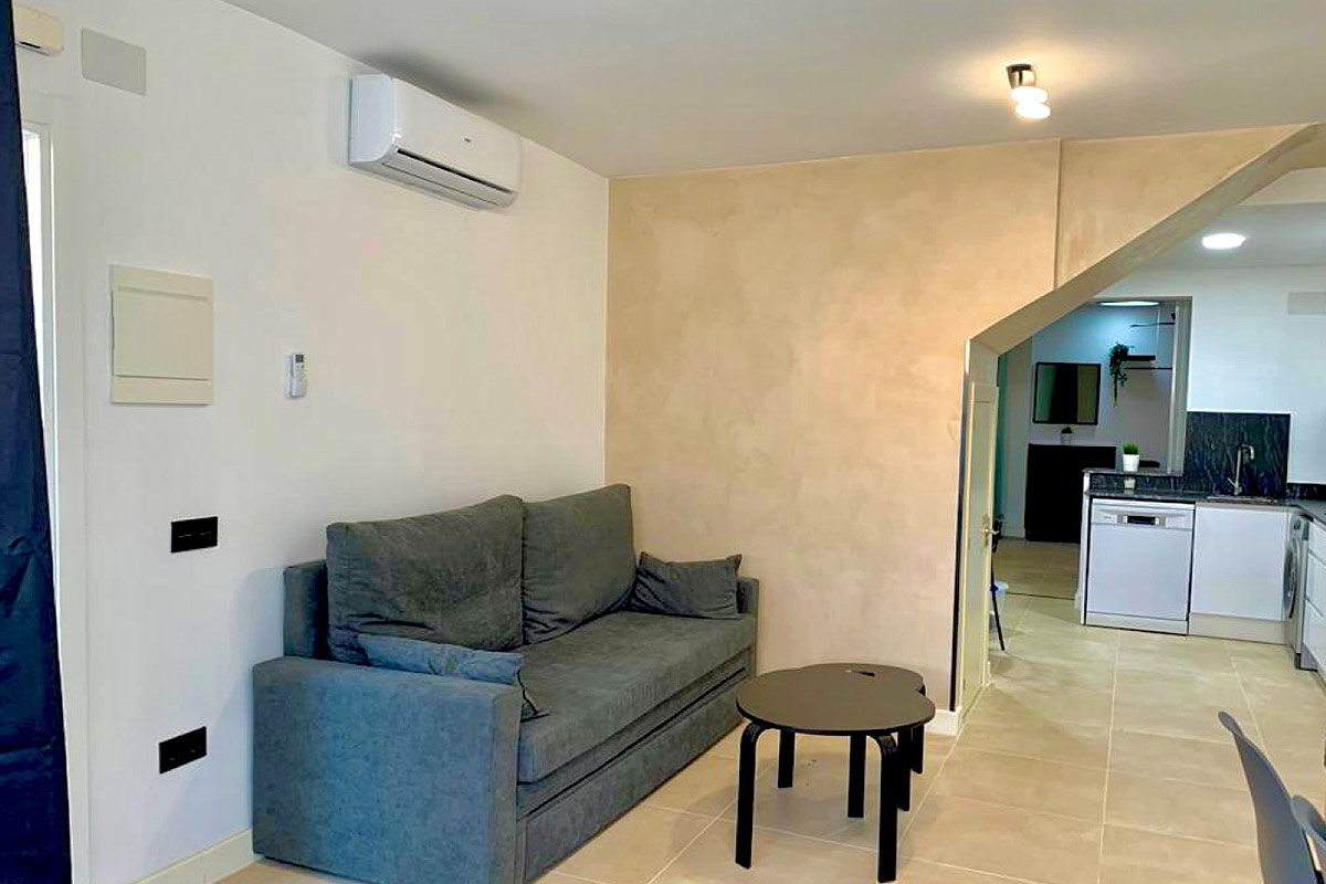 Sofá gris en salón moderno junto a pared beige y vista parcial de la cocina abierta.