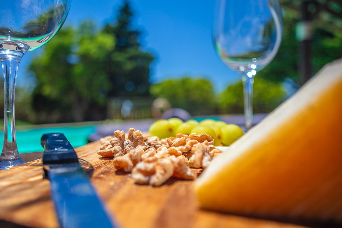 Detalle de nueces, uvas y queso sobre tabla de madera en primer plano, con copas de vino vacías y piscina del Chalet La Ahumada al fondo.