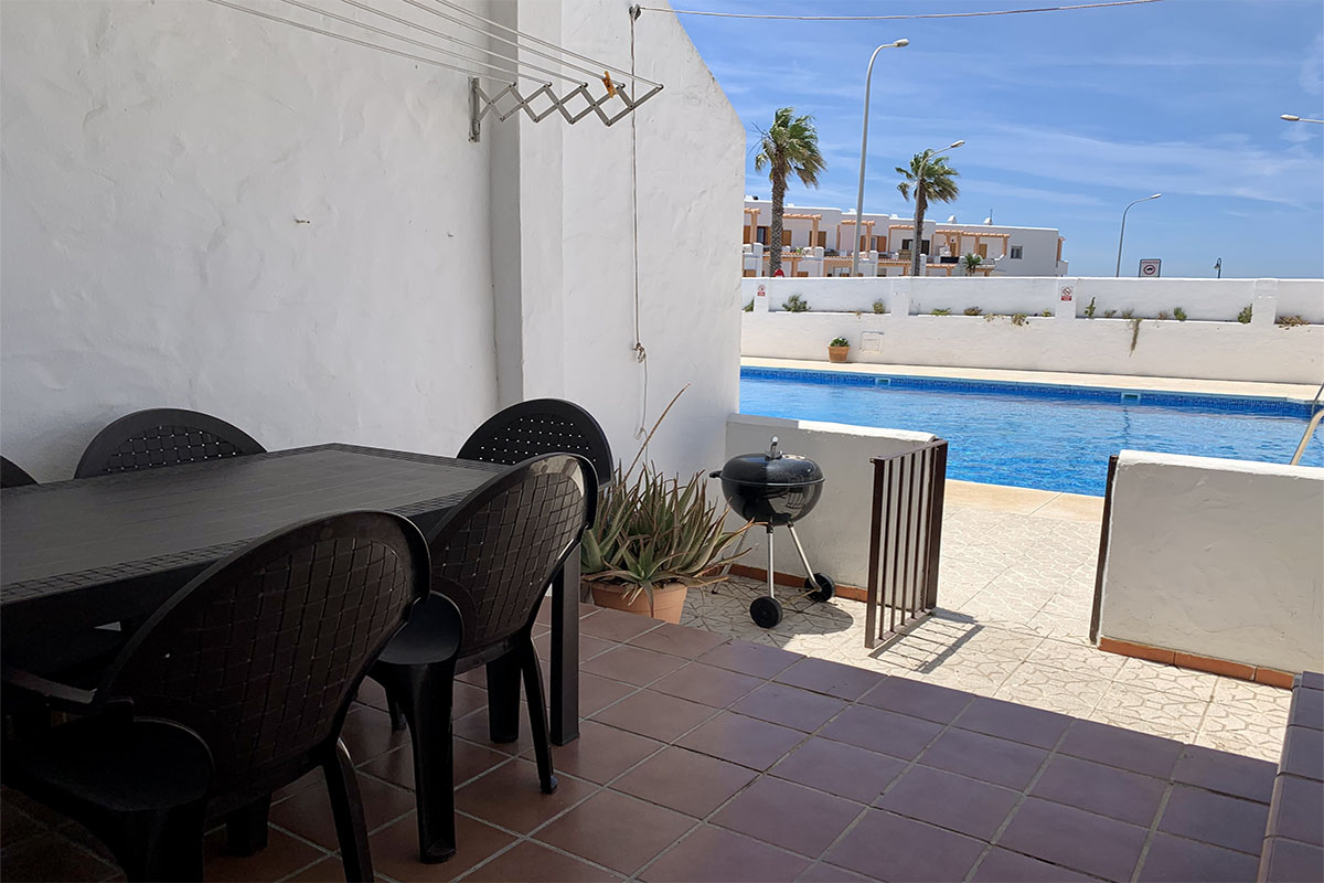 Comedor exterior con mesa negra y sillas, junto a piscina comunitaria con barbacoa.