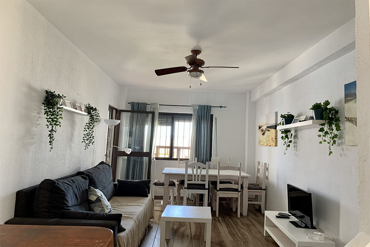 Sala de estar con sofá oscuro, lámpara blanca, TV y una cocina abierta moderna al fondo.