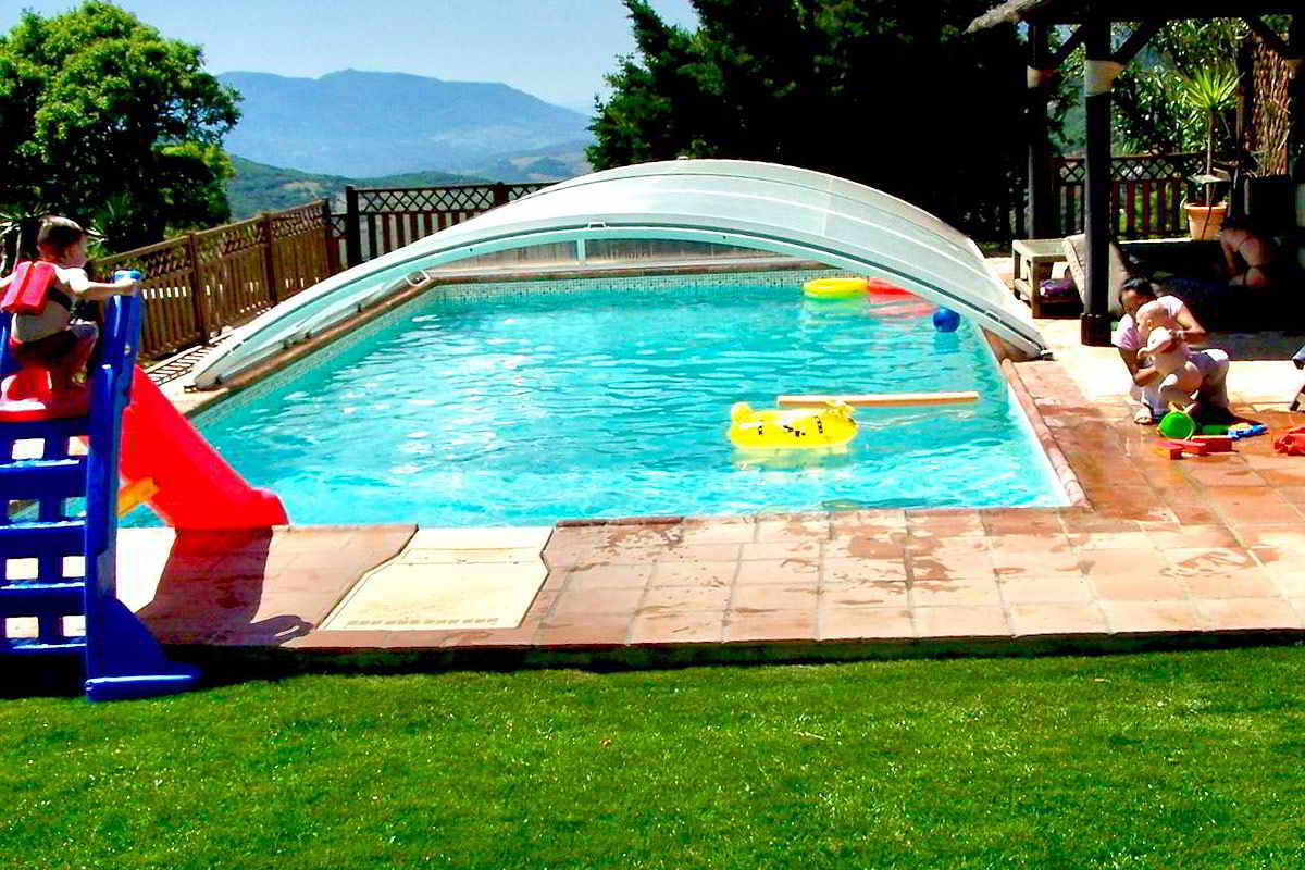 Piscina exterior de Villa Paraíso en el chalet La Ahumada con cubierta móvil, rodeada de césped y zona de juegos infantiles, con vistas a la sierra de Tarifa.