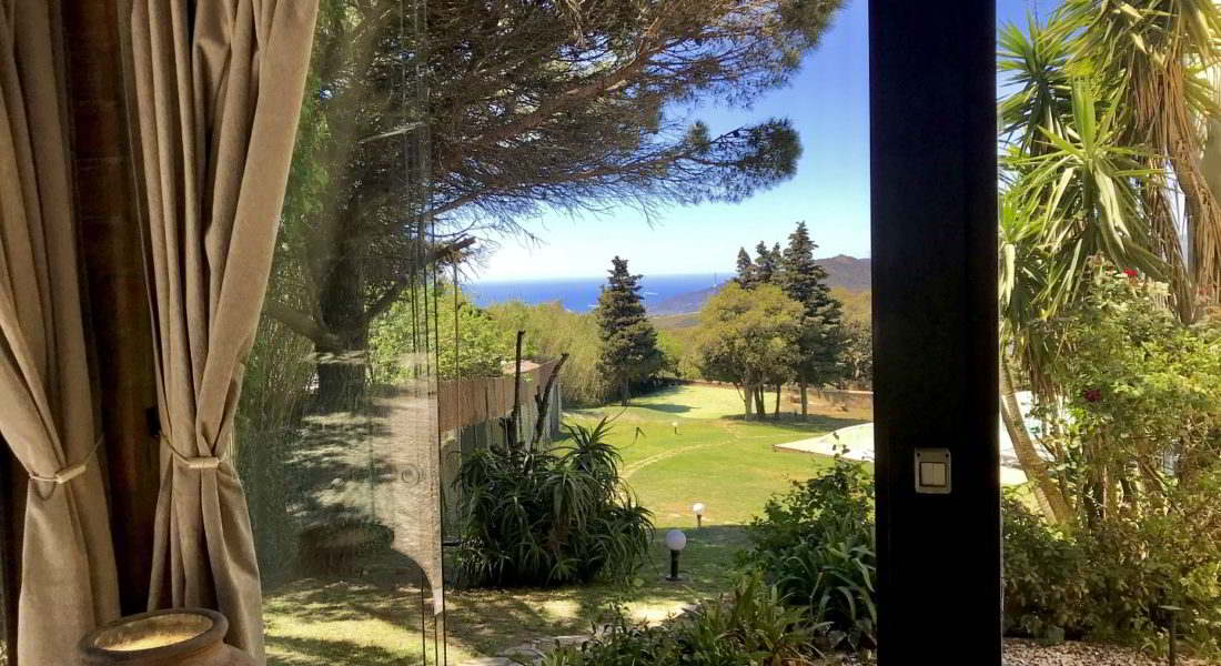 Vista desde el interior del chalet hacia el jardín con árboles, césped y el mar al fondo en un entorno natural.