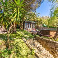 Zona ajardinada escalonada con camino de piedra, piscina elevada de madera, hamaca atada entre árboles y una pequeña cabaña de madera al fondo rodeada de palmeras y vegetación.