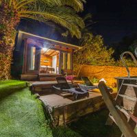 Vista nocturna de una acogedora cabaña de madera con puertas abiertas, rodeada de palmeras y vegetación, junto a una piscina elevada de madera y tumbonas iluminadas cálidamente sobre una tarima.