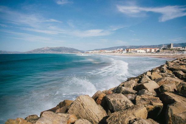 Vista de la playa de Tarifa con olas rompiendo contra las rocas y el pueblo al fondo.