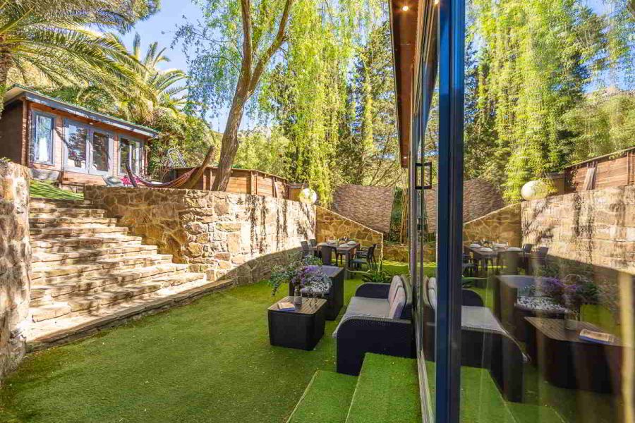 Vista desde la esquina del jardín de la casa Nepal en Villas La Ahumada, con escalera de piedra, salón acristalado reflejando la vegetación, zona chill out con sillones y mesa, y al fondo el comedor exterior bajo árboles frondosos