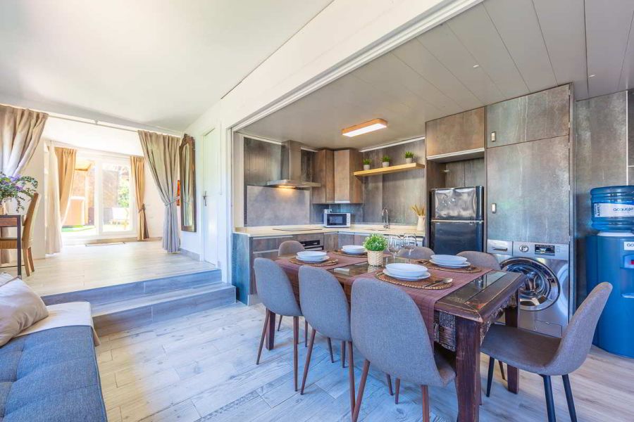 Cocina moderna con acabados de piedra y madera, equipada con electrodomésticos como horno, lavadora y frigorífico, mesa de comedor con vajilla colocada, escaleras que conectan con zona luminosa de acceso al jardín.