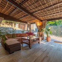 Porche cerrado del chalet La Ahumada con techo de cañizo, cortinas recogidas, sofá chaise longue de terciopelo marrón y mesa de centro de madera, rodeado de vegetación y luz natural.
