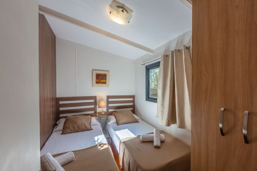 Dormitorio con dos camas individuales en la casa Nepal de Villas La Ahumada, con cabeceros de madera, lámpara de noche encendida, ventana con cortinas claras, armario de madera, decoración minimalista y luz natural