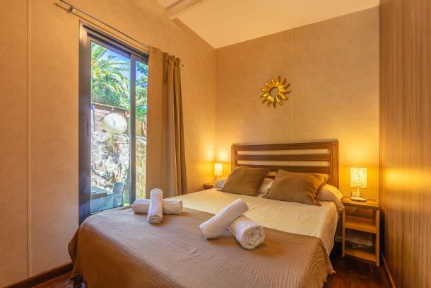Dormitorio principal de la casa Nepal en Villas La Ahumada, con cama doble vestida en tonos tierra, cabecero de madera, lámparas de noche encendidas, decoración en forma de sol dorado en la pared y ventanal con vistas al jardín exterior