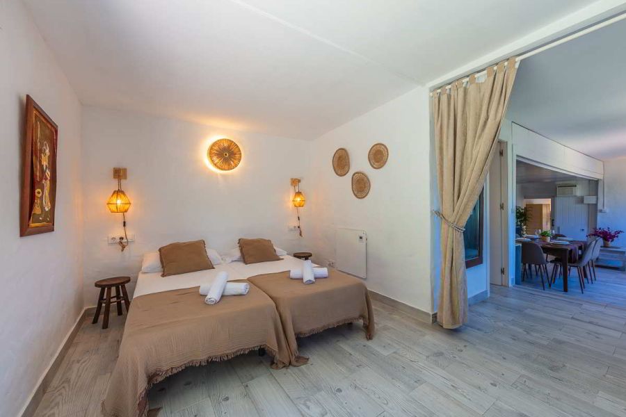 Dormitorio doble de la Villa Verde con dos camas individuales unidas, colchas en tonos tierra, lámparas colgantes artesanales, decoración étnica en pared blanca y cortina beige que comunica con la cocina y salón.