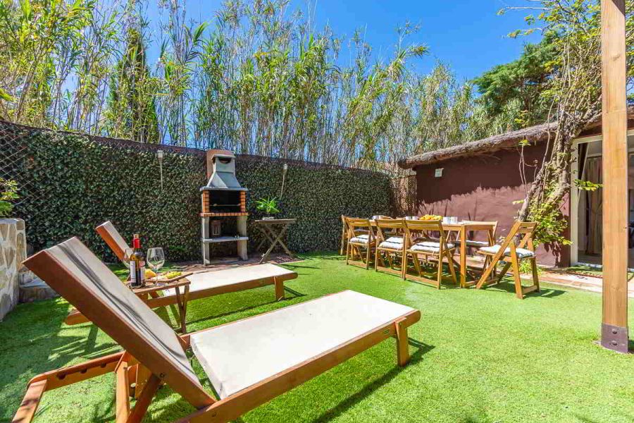 Jardín soleado de Villa Verde con dos tumbonas en primer plano, una mesa larga preparada para comer y una barbacoa de obra al fondo, todo rodeado de vegetación.