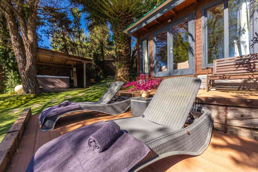Primer plano de dos tumbonas de ratán con toallas moradas en una tarima de madera, situadas frente a una cabaña de madera con puertas de cristal, junto a una jardinera con flores fucsias y un banco de madera; al fondo, un jacuzzi cubierto entre árboles.