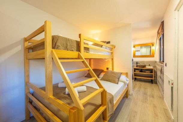 Cama litera de madera con colchón doble en la parte inferior y colchón individual en la parte superior, con ropa de cama en tonos marrones y almohadas decorativas, vista parcial del baño con lavamanos de piedra y espejo de madera al fondo.