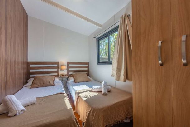 Dormitorio con dos camas individuales en la casa Nepal de Villas La Ahumada, con cabeceros de madera, lámpara de noche, ventana con vistas al exterior ajardinado, armario y ropa de cama en tonos tierra