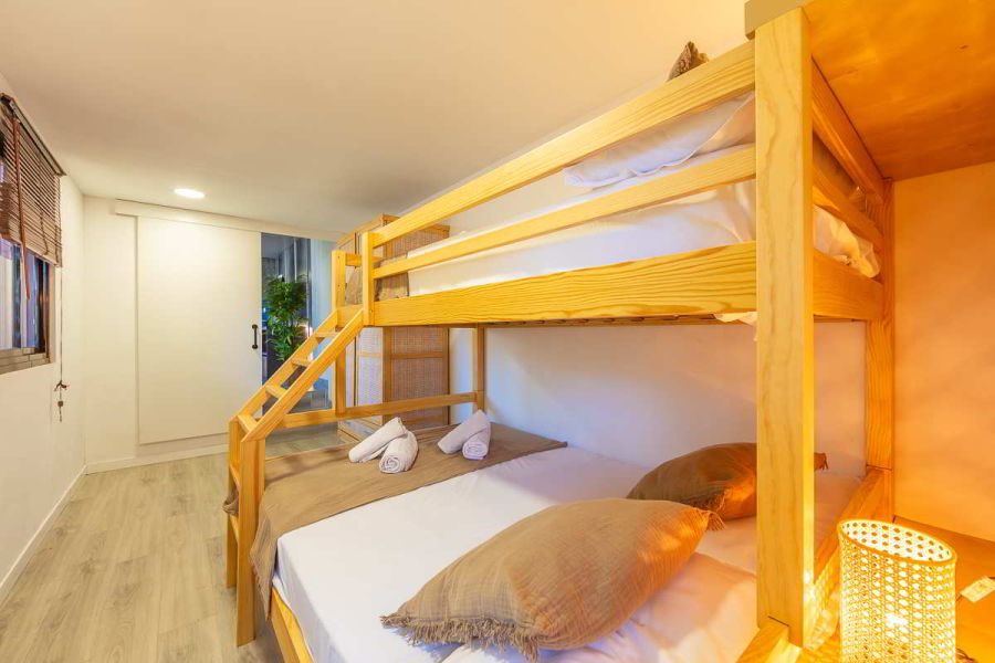 Detalle de una litera de madera con cama doble inferior y cama individual superior, ropa de cama en blanco y marrón, cojines decorativos, lámpara de mimbre encendida y puerta corredera blanca al fondo junto a planta decorativa