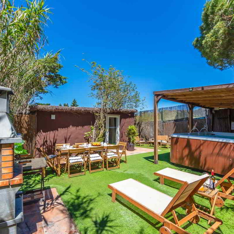 Vista del jardín privado de la Villa Verde en Tarifa, con césped artificial, tumbonas de madera, una mesa grande de comedor al aire libre, barbacoa de obra y un jacuzzi bajo pérgola, todo rodeado de vegetación natural.