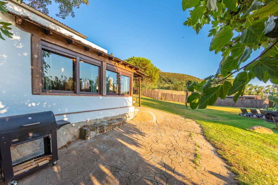 Terraza lateral de Villa Paraíso en Villas La Ahumada, Tarifa, con ventanales de madera, barbacoa, amplio jardín y tumbonas bajo un cielo despejado.