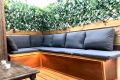 Banco de madera con cojines grises en una terraza rodeada de plantas y enredaderas verdes.