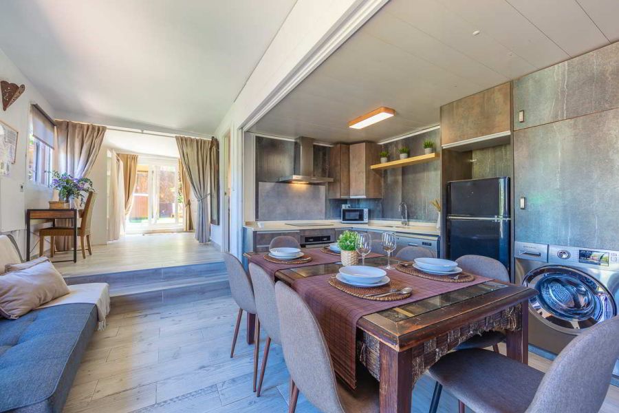 Vista de la cocina abierta de la Villa Verde con mobiliario en tonos cobre envejecido, mesa de comedor rústica con vajilla dispuesta, lavadora, nevera negra, plantas decorativas y fondo con salida a la terraza.