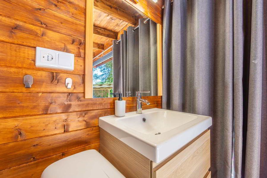 Detalle del lavabo moderno con mueble de madera clara y grifería cromada sobre pared de madera natural, con espejo rectangular y cortinas grises que cubren la entrada de luz exterior, creando un ambiente cálido y privado