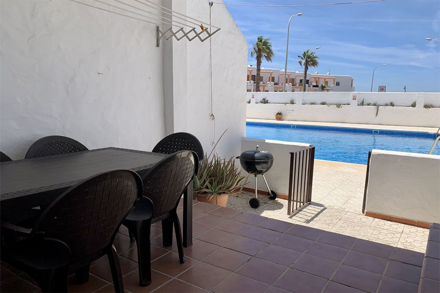 Comedor exterior con mesa negra y sillas, junto a piscina comunitaria con barbacoa.