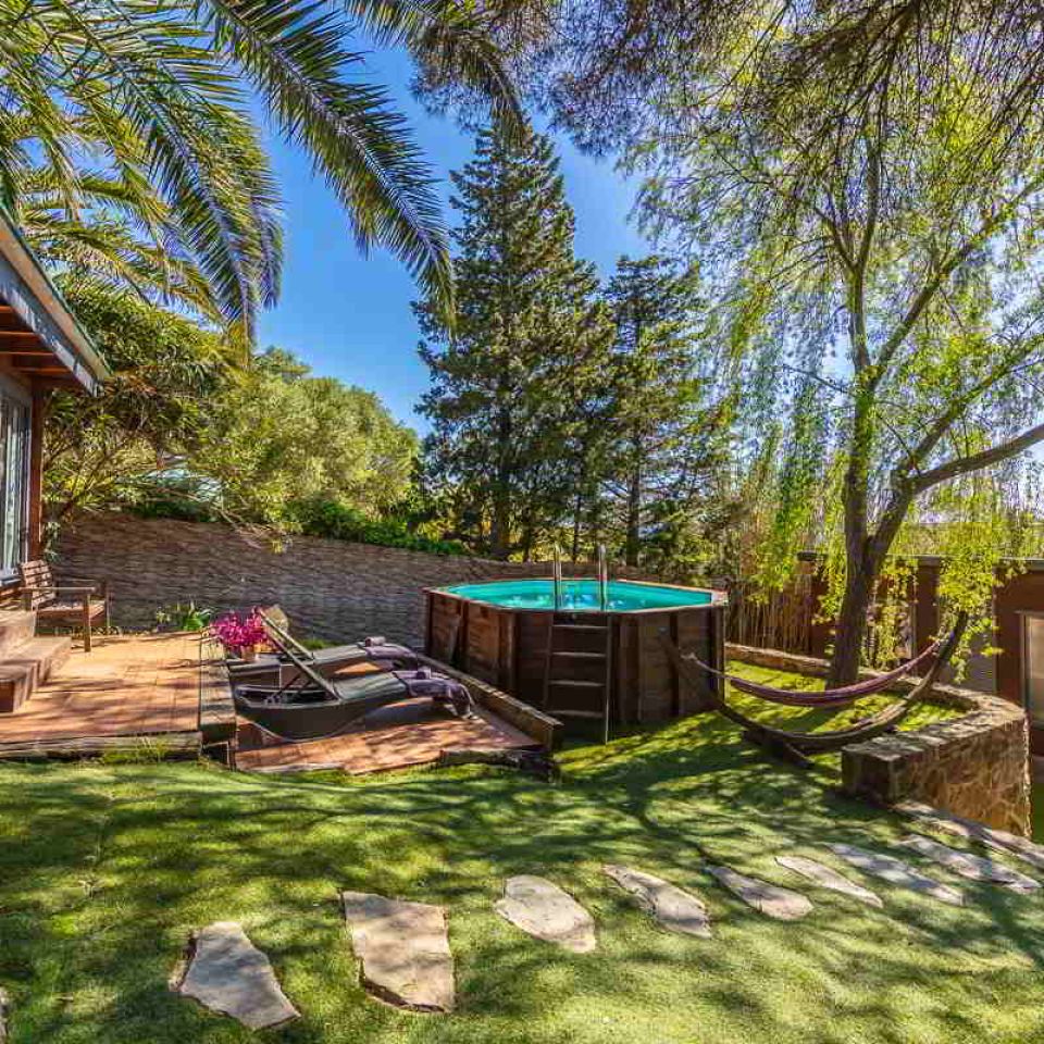 Imagen del jardín con piscina elevada de madera, tumbonas sobre tarima, una hamaca entre árboles y una cabaña de madera bajo las palmeras en el alojamiento en Tarifa.