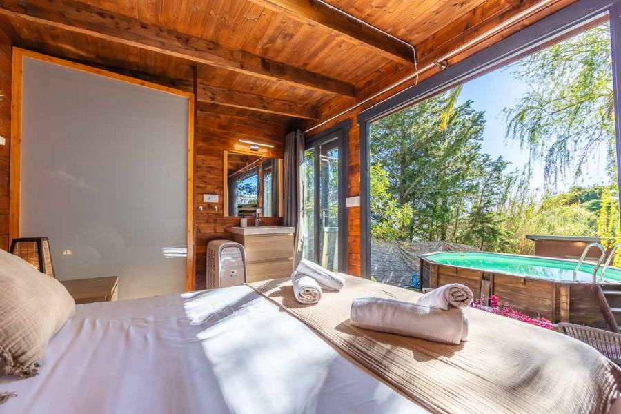 Vista desde la cama de una habitación de madera con techos de vigas, toallas enrolladas sobre una manta beige, lavabo con espejo y mueble claro, mampara translúcida a la izquierda, grandes ventanales abiertos que dan acceso directo a una terraza con piscina elevada rodeada de naturaleza.