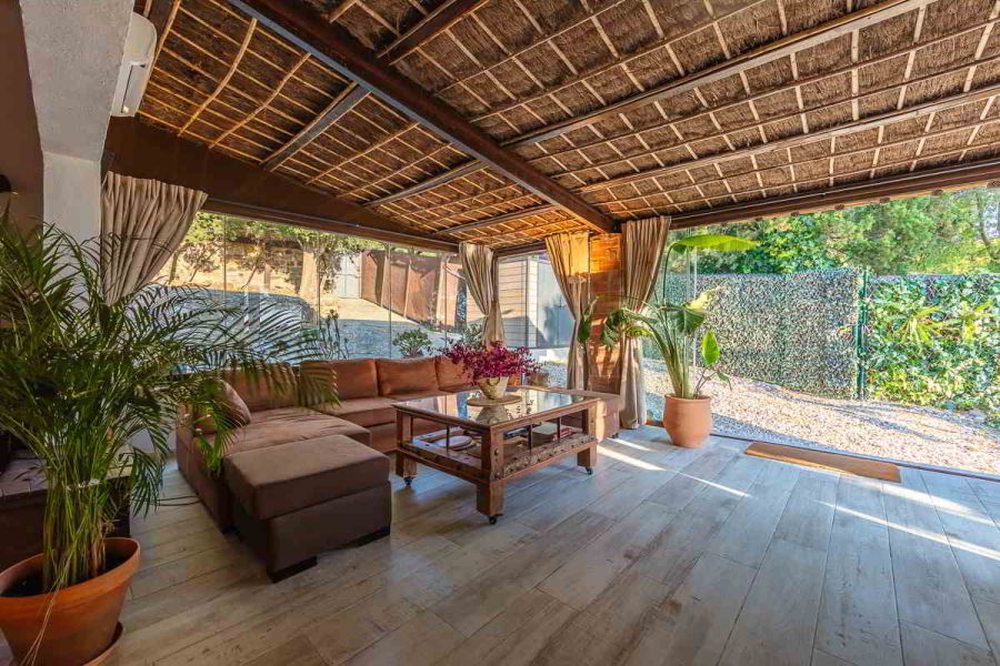 Porche cerrado del chalet La Ahumada con techo de cañizo, cortinas recogidas, sofá chaise longue de terciopelo marrón y mesa de centro de madera, rodeado de vegetación y luz natural.