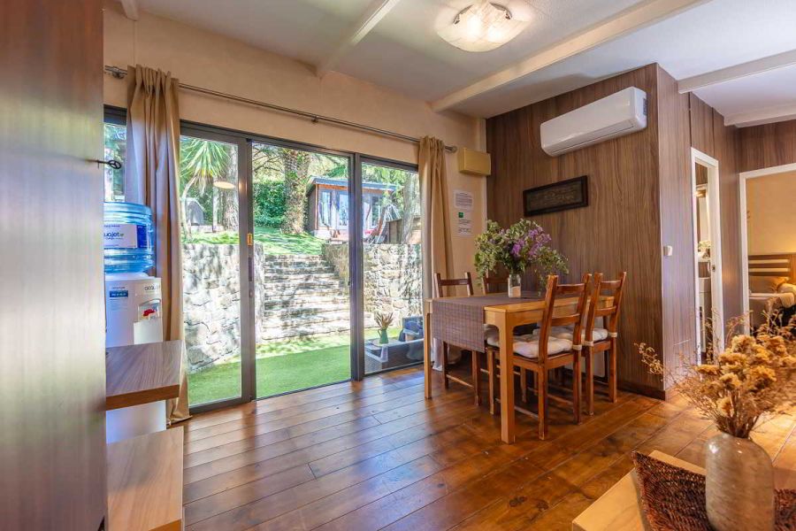 Comedor de la casa Nepal en Villas La Ahumada, con mesa de madera decorada con flores, suelo de madera natural, dispensador de agua a la izquierda y grandes ventanales con vistas al jardín escalonado y la casita exterior entre árboles