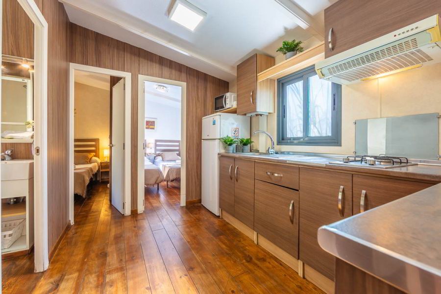 Cocina de la casa Nepal en Villas La Ahumada, con muebles de madera, encimera de acero, ventana con luz natural, frigorífico, microondas y acceso a dos dormitorios con camas individuales y baño lateral