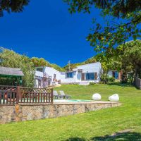 Vista del jardín del Chalet La Ahumada con piscina vallada, pérgola de sombra, tumbonas y fachada blanca del edificio principal al fondo.